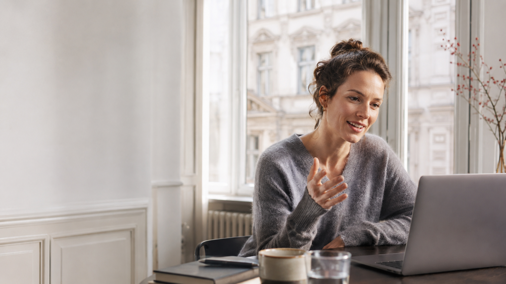 Woman in a grey jumper gesturing while she talks to her laptop